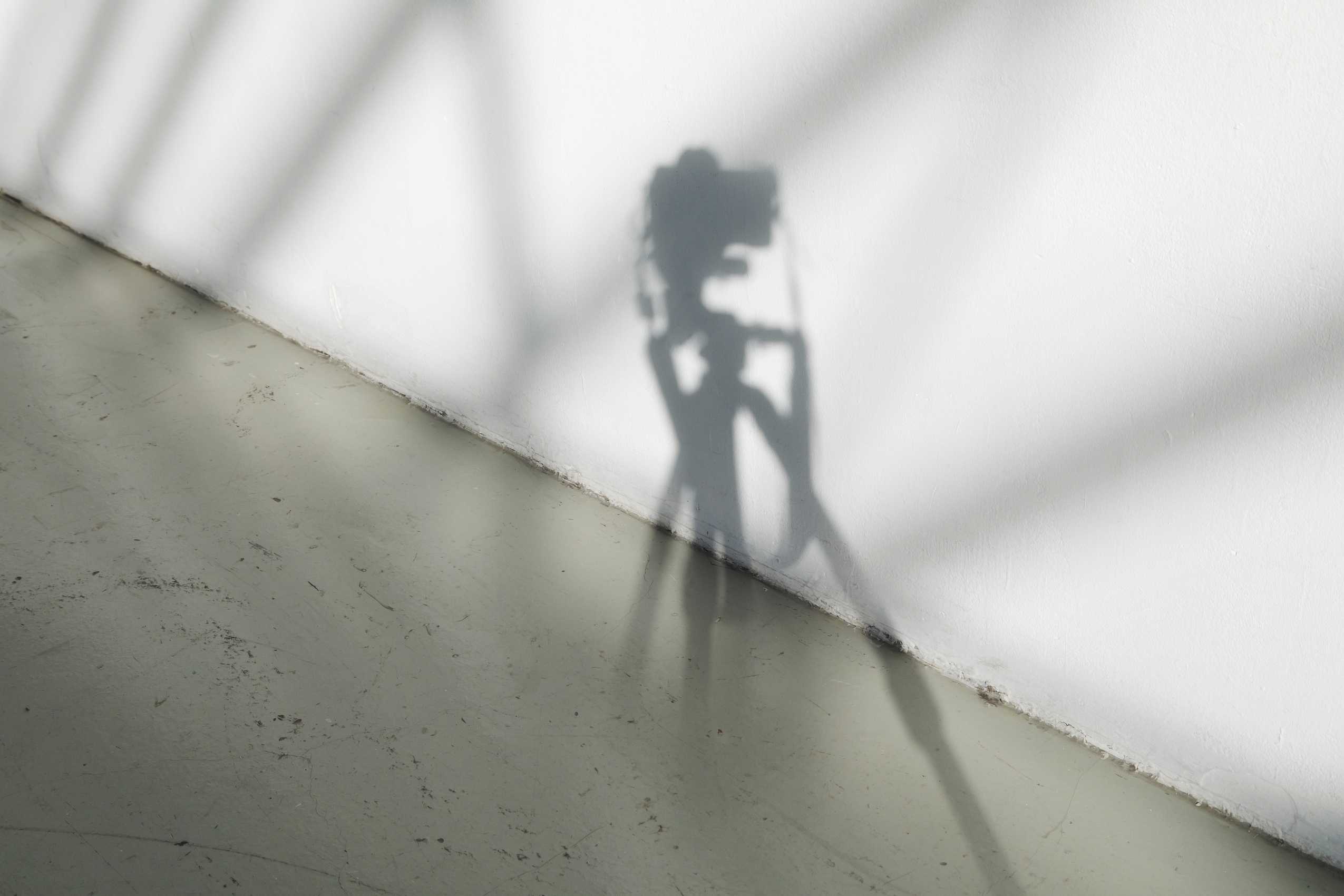 À l’intersection du sol gris et du mur blanc, l’ombre d’un appareil photo se reflète, flouté par la lumière.