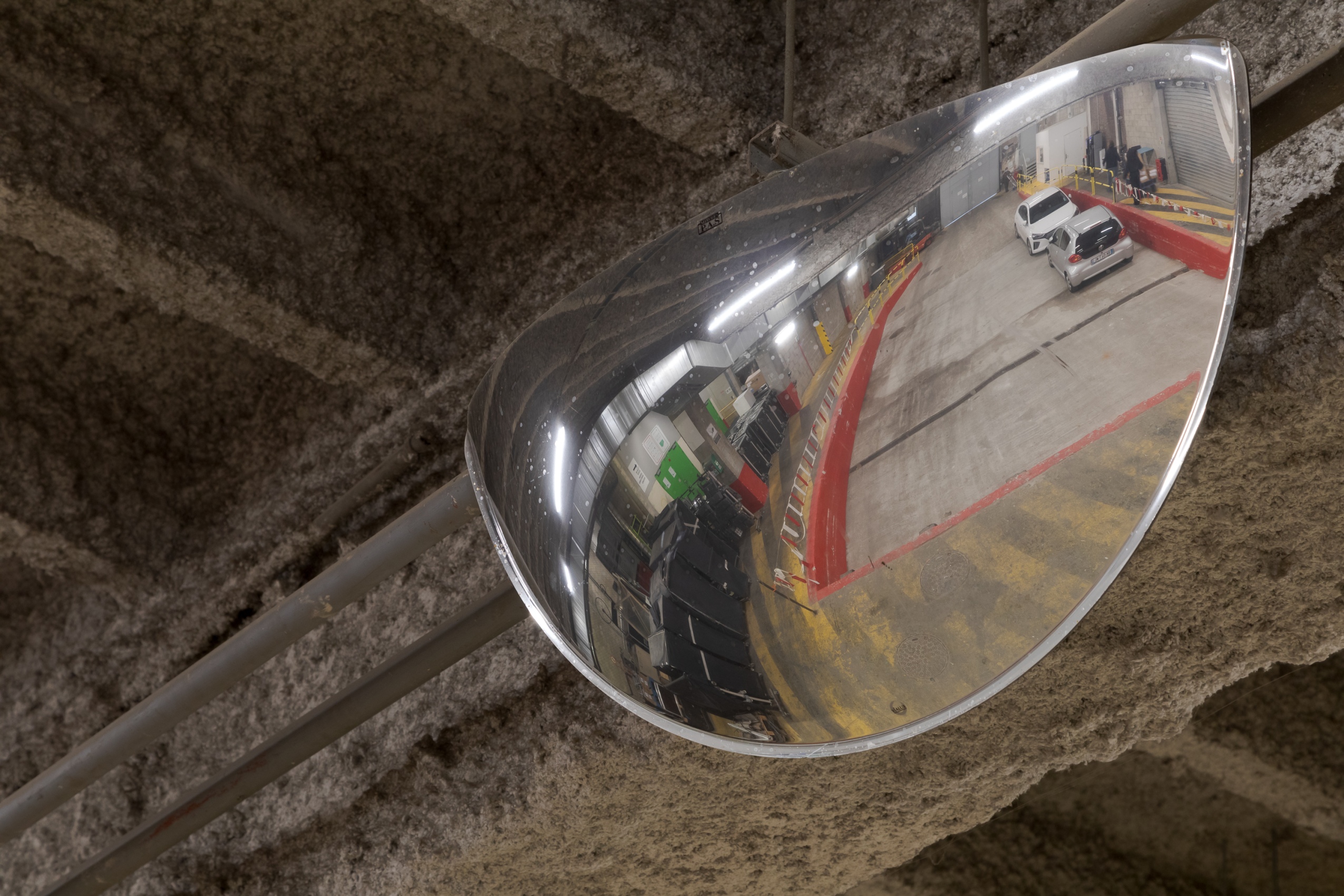 Un miroir courbe en métal est suspendu aux poutres floquées du quai de déchargement : on aperçoit dans le miroirs les lignes jaunes de la signalétique au sol ainsi que plusieurs voitures garées. 