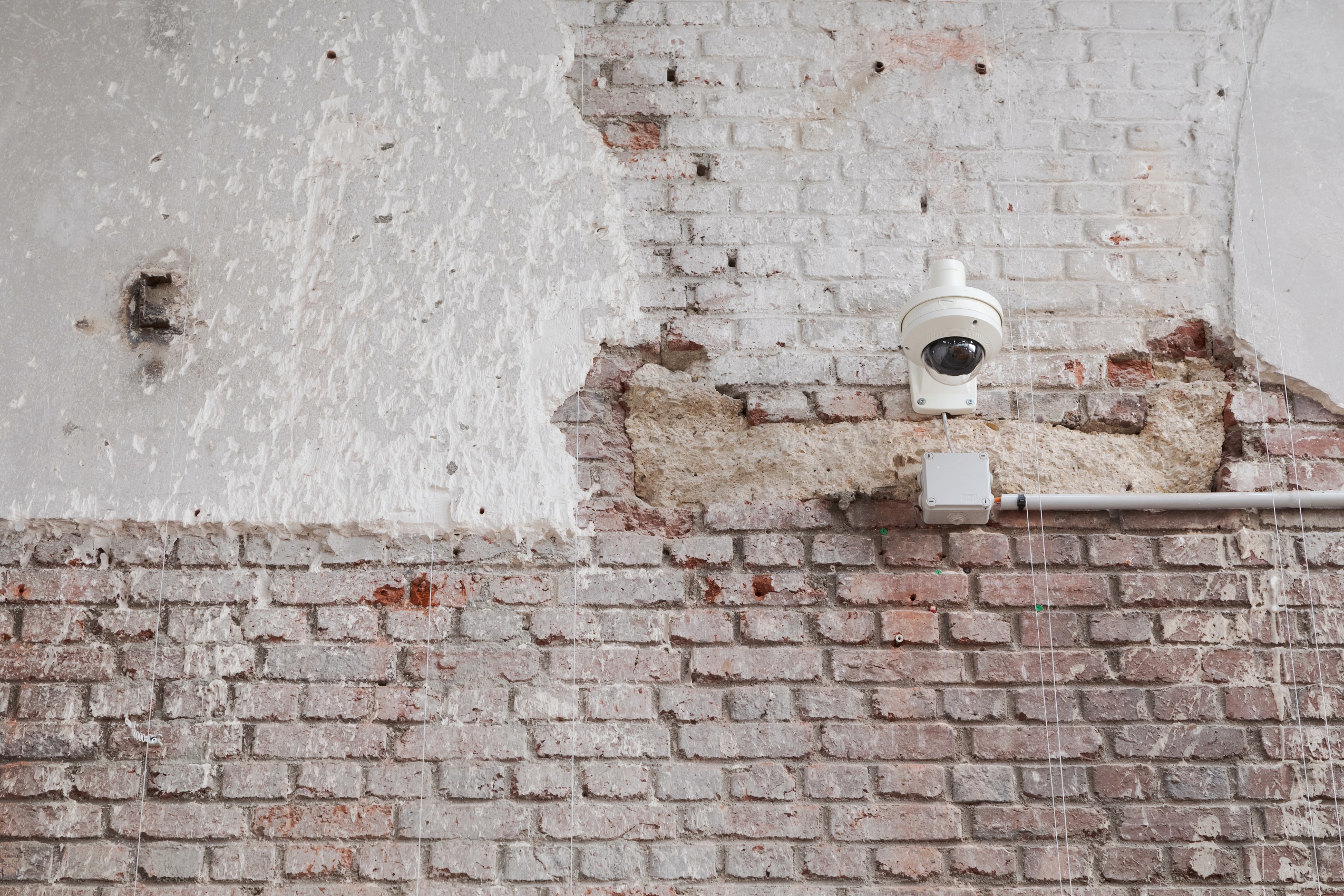 Sur un mur de briques rouges ternies par le temps, une caméra de surveillance blanche semblable à un petit lampadaire se détache. 