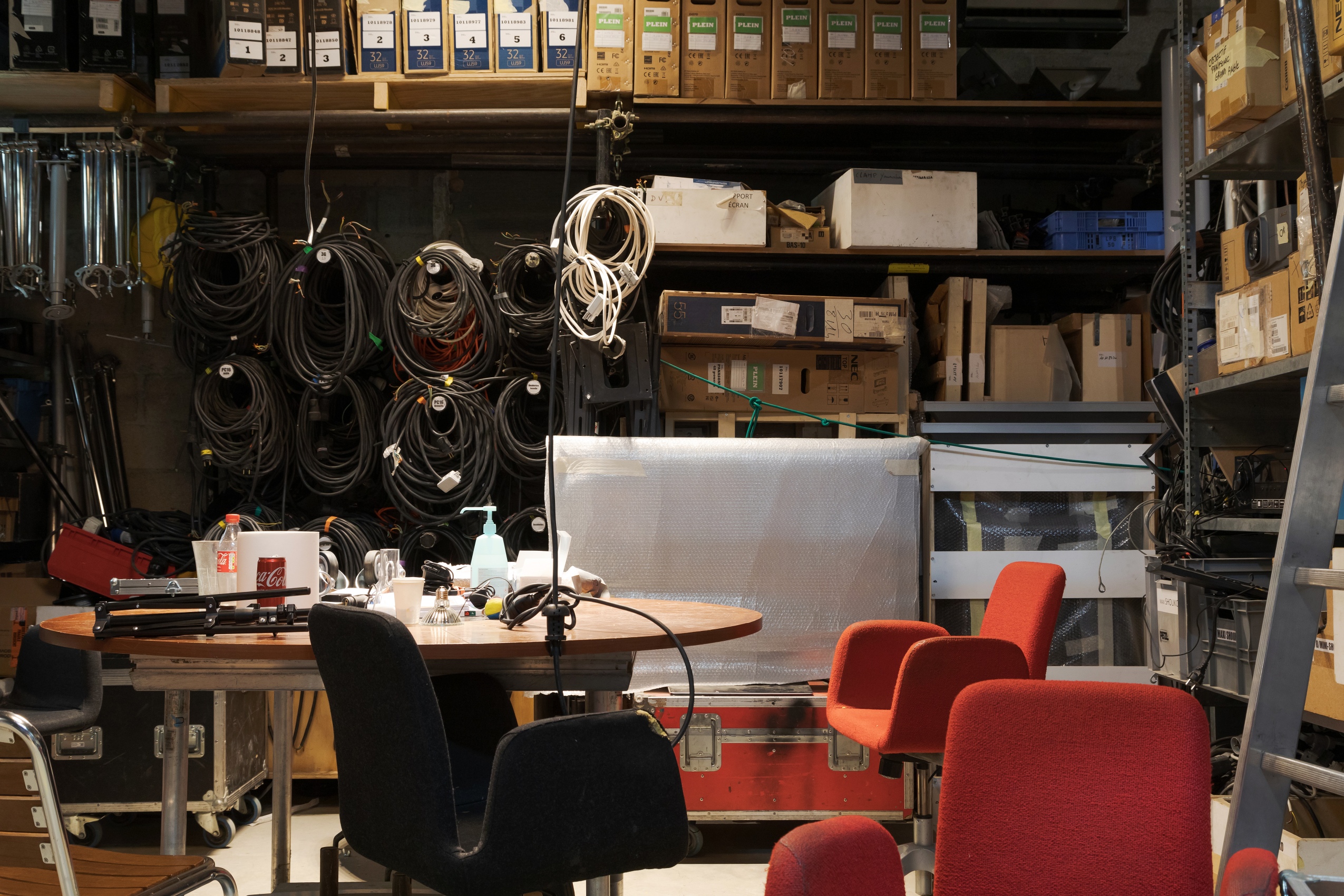 L’atelier de la régie audiovisuelle est organisé autour d’une table centrale, entouré de fauteuils rouges et noirs. Les murs sont couverts de câbles, de projecteurs, de rouleaux de plastique coloré, d’échelles ou de cartons.  
