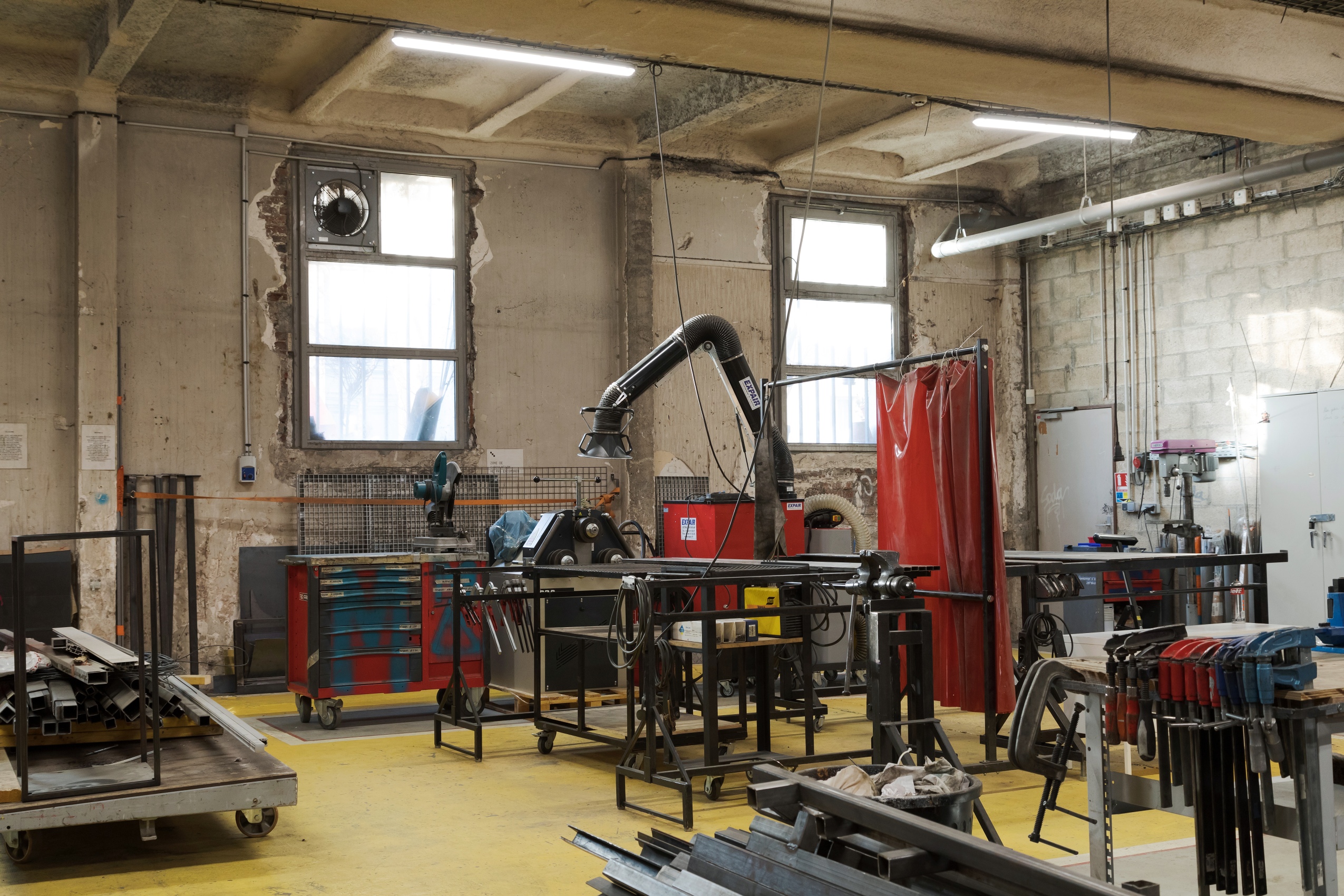Dans un grand espace au sol jaune, de nombreuses machines et outils sont soigneusement rangées : étaux, scie circulaire ou profilés en acier. 